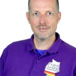 Ein Porträt von Kai Bölle. Ein Mann mit kurzen Haaren und Bart steht vor einem weißen Hintergrund. Er trägt ein lila Poloshirt mit dem Logo des CSD Deutschland e.V. auf der linken Seite.